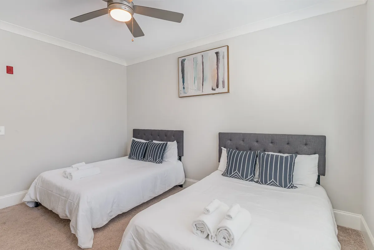 A cozy bedroom with two beds featuring tufted headboards, striped pillows, and framed wall art. A ceiling fan above ensures comfort, and ample natural light enhances the inviting mood.