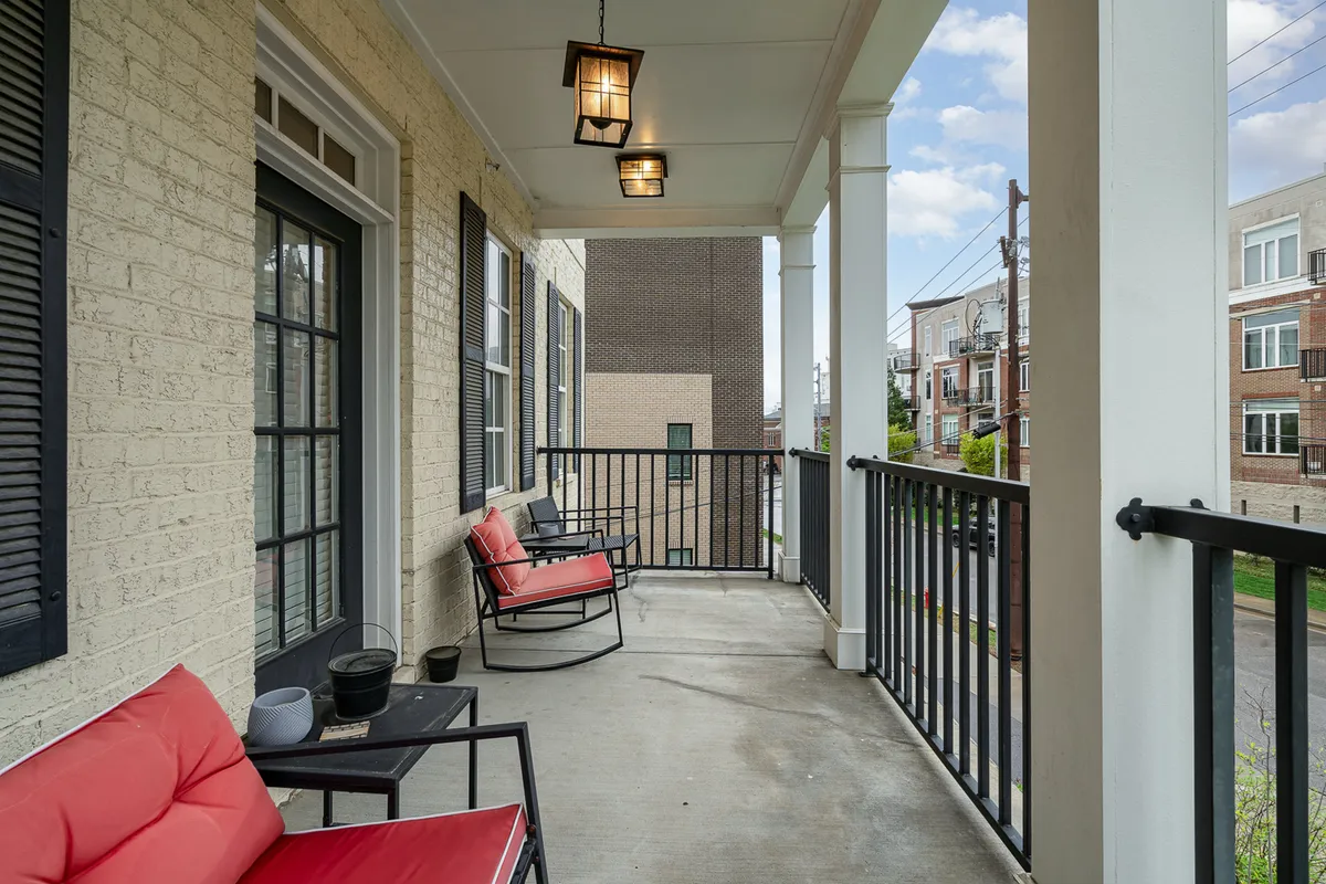 An exterior view of a porch with red chairs, black railing, hanging lanterns, and a brick facade. The setup suggests a comfortable spot for relaxing, reading, or sharing coffee, with an emphasis on outdoor living and southern charm.