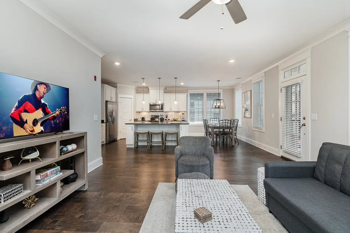 An inviting, open-concept interior with a plush sofa facing a mounted smart TV, a spacious kitchen island with seating, and a dining table set for guests. Large windows flood the living space with natural light, making it perfect for socializing, movie nights, or casual dining.