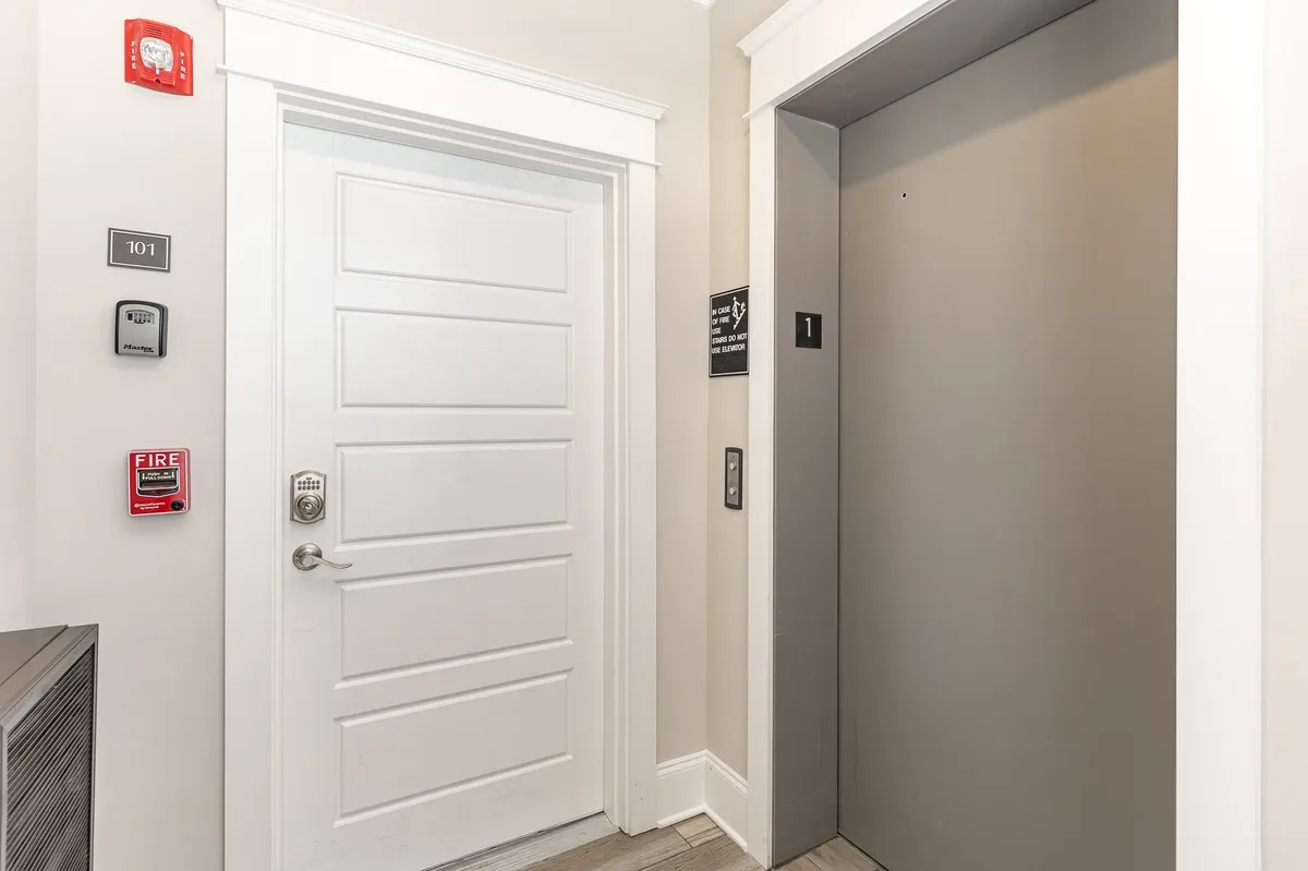 An interior shot of a white entry door adjacent to an elevator, featuring number signage and a fire alarm. The scene is clean and functional, evoking the smooth transition from the garage or street into the living areas.