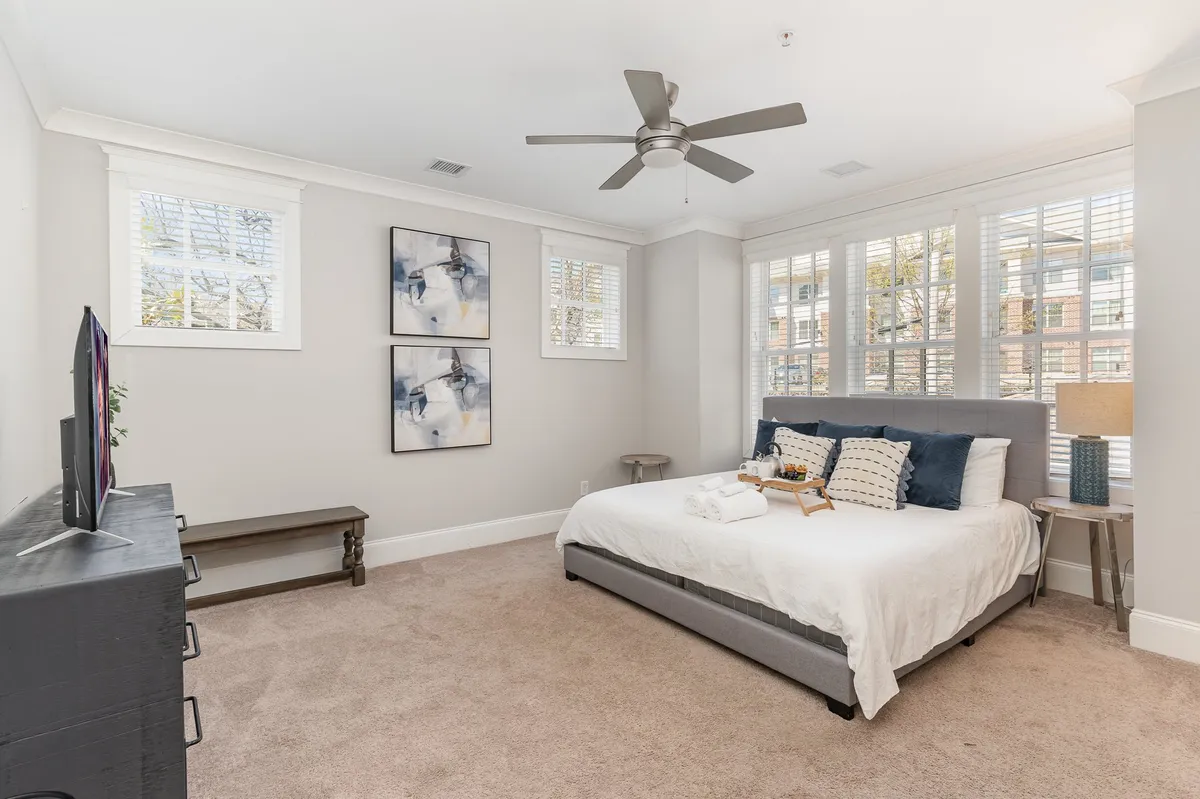 A serene bedroom scene with a neatly made bed, soft pillows, and abstract art adorning the walls. A wooden bedside table sits next to the bed, and natural light filters into the room, creating a tranquil and restful atmosphere.