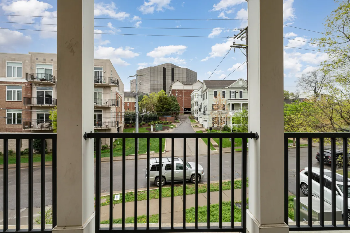 A lively view from a balcony overlooking surrounding buildings and a green space below. The image suggests a prime spot for enjoying morning coffee, sunsets, or recapping the day with friends while taking in the local vibe.